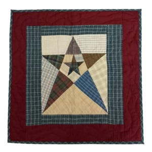 May include: A square quilted wall hanging with a star design. The star is composed of various fabric patterns in beige, blue, green, and brown. It is centered on a cream-colored background, framed by a green plaid border and a deep red outer border.
