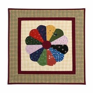 May include: A square, handmade quilt with a floral design. The center features a large, multi-colored flower composed of various patterned fabrics. The flower is surrounded by a burgundy border and a plaid outer border.