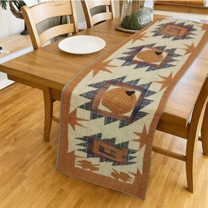 May include: A rectangular table runner with a cream background and a repeating pattern of geometric shapes and pumpkin-like designs in shades of orange, brown, and blue. The runner is placed on a wooden table.