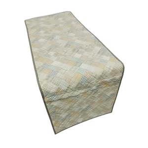 Puede incluir: Camino de mesa acolchado rectangular con diseño de patchwork. El camino de mesa presenta una mezcla de colores neutros, incluyendo beige, crema y azul claro, con un sutil estampado de cuadros. Los bordes están acabados con un ribete gris oscuro.