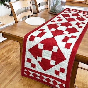 May include: A red and white quilted table runner with a geometric pattern. The runner is rectangular and features a repeating design of red squares and diamonds on a white background. The runner is placed on a wooden table.