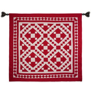 May include: A red and white quilt with a geometric pattern. The quilt is made of squares and diamonds, and it has a border of small diamonds. The quilt is hanging on a wall.