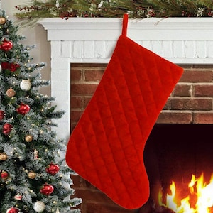 Op de afbeelding: Een rode, gewatteerde kerstsok die boven een open haard hangt. De sok is gemaakt van een zachte, pluche stof en heeft een traditioneel ontwerp.