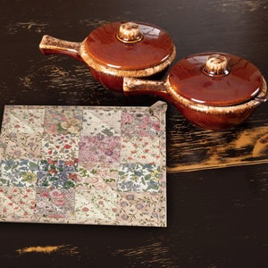 May include: Two brown ceramic casserole dishes with lids and handles on a dark wood surface. A floral patchwork fabric hot pad is in the foreground.