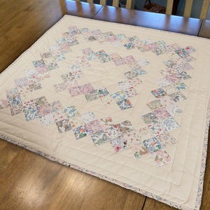 Könnte beinhalten: Ein weißer gesteppter Tischläufer mit einem Rautenmuster aus floralen Stoffquadraten. Der Läufer hat einen rosa-weißen Rand.