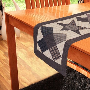 Peut inclure: Un chemin de table matelassé brun et blanc avec un grand motif d'étoile. Le chemin de table est fait de tissu patchwork et a une bordure brun foncé.