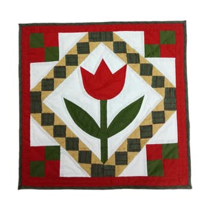May include: A square quilt with a central red tulip design. The tulip is set against a white background, surrounded by green leaves. The quilt features a red border and accents of green, white, and gold checkered patterns.