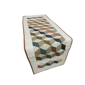 May include: A quilted table runner featuring a geometric cube design in various plaid and solid fabrics. The runner has a cream-colored background with a brown border. The cube pattern includes shades of red, green, blue, and brown.