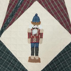 May include: A quilted Christmas-themed design featuring a nutcracker figure. The nutcracker is dressed in a red and blue uniform with a white beard and mustache. The background is a patchwork of plaid and patterned fabrics.