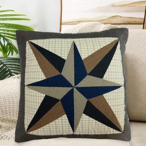 Puede incluir: Cojín decorativo cuadrado con diseño de estrella. La estrella presenta triángulos azul marino, marrón y negros sobre un fondo crema y a cuadros. El cojín tiene un borde gris oscuro.