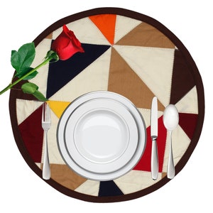 May include: A white dinner plate with a smaller white plate inside, a silver fork, a silver knife, and a silver spoon on a round, multi-coloured patchwork placemat with a red rose on the left side.