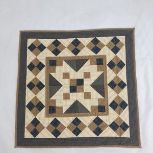 Könnte beinhalten: Ein quadratischer Patchwork-Quilt mit einem zentralen Sternmotiv aus cremefarbenen, beigen und dunkelblauen karierten Stoffquadraten. Das Design umfasst eine Bordüre aus abwechselnden rautenförmigen Patches und einen dunkelgrauen Außenrahmen.