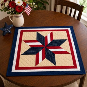 May include: A square quilted table mat with a patriotic star design. The star is composed of navy blue dotted fabric, red, and white stripes, set against a cream background. The mat is bordered by red, white, and navy blue.