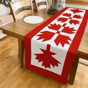May include: A red and white table runner featuring a repeating pattern of stylized maple leaves. The runner is rectangular and placed on a wooden table, adding a decorative touch to the dining area.