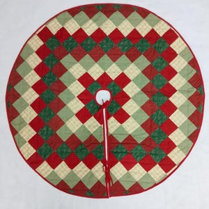 May include: A round Christmas tree skirt with a quilted pattern. The skirt features a patchwork design of red, green, and cream-colored squares. A red border and ties complete the festive look. The skirt measures approximately 50 inches in diameter.