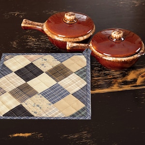 Puede incluir: Un posavasos acolchado con un borde azul y un diseño de patchwork de cuadrados beige, marrón y azul. Dos platos de cerámica marrón con tapas están sobre una superficie de madera oscura.