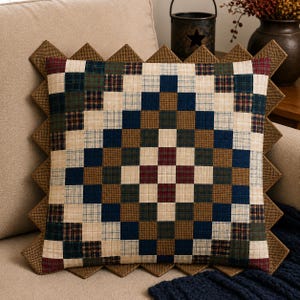 May include: A square patchwork cushion with a geometric pattern in shades of blue, green, brown, and cream. The cushion has a decorative, zig-zag edge in a complementary brown fabric. The cushion is placed on a beige sofa.