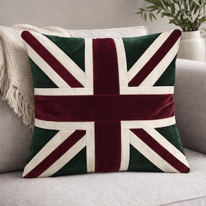 May include: Square cushion featuring the Union Jack flag design. The cushion has a dark green background with a burgundy cross and diagonal stripes, and off-white stripes. The cushion is placed on a grey sofa.