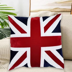 May include: Square decorative pillow featuring the Union Jack, the national flag of the United Kingdom. The pillow has a navy blue background with red and white design elements. The pillow is on a beige couch.