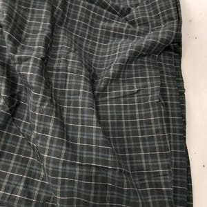May include: Dark green plaid fabric with white and light blue lines. The fabric is folded and draped, creating shadows and texture. The pattern is a classic grid design, suitable for apparel or home decor.