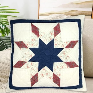 May include: Square throw pillow with a quilted star design. The star features a navy blue center and red and floral print accents on a white background. The pillow has a navy blue border.