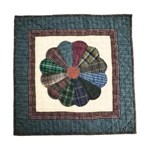May include: A quilted square with a floral design. The center features a flower made of plaid fabric petals in various colors, including green, red, and blue. The flower is set against a cream-colored background, framed by a plaid border and a dark teal outer border.