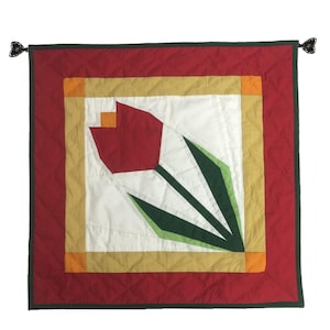 May include: A red and green quilted wall hanging with a geometric tulip design. The tulip is surrounded by a gold and orange frame.