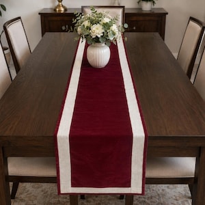 May include: A rectangular table runner featuring a deep burgundy velvet centre panel bordered by a light beige linen-textured fabric. The runner is centred on a dark brown wooden dining table with matching chairs and a white vase of flowers.