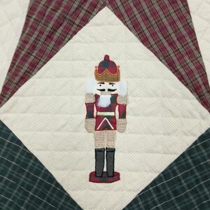 May include: A quilted Christmas decoration featuring a nutcracker design. The nutcracker is in the center, with a red and white uniform, black boots, and a gold crown. The background is a patchwork of cream, red, and green plaid fabrics.