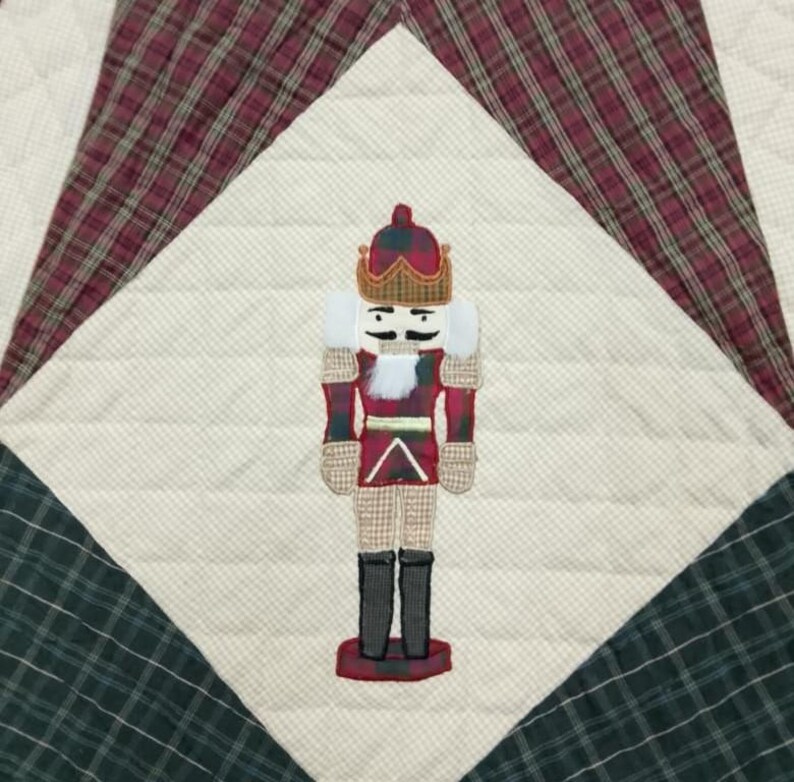 May include: A quilted Christmas decoration featuring a nutcracker design. The nutcracker is in the center, with a red and white uniform, black boots, and a gold crown. The background is a patchwork of cream, red, and green plaid fabrics.
