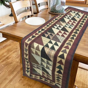 May include: A quilted table runner with a geometric pattern of brown, green, and beige triangles. The runner has a burgundy border with a smaller triangle pattern.