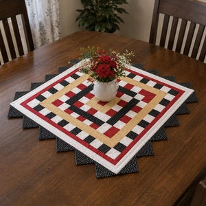 May include: A square, quilted table runner with a geometric pattern in red, black, white, and gold. The runner has a zigzag edge and a small white vase with red flowers in the centre. The table is made of dark wood.