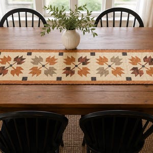 May include: A rectangular table runner featuring a repeating quilt-style pattern of stylized leaves in shades of orange, brown, and grey on a cream background, bordered by an orange fabric with small dark blue triangles at each end.