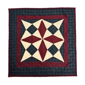 May include: Square quilt with a central starburst design in cream, burgundy, and dark green. The starburst is surrounded by a burgundy border and a dark green plaid border. The quilt is likely made of cotton or a similar fabric.
