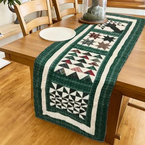 May include: A green and white quilted table runner with a geometric pattern. The runner features a central design of stars, triangles, and other shapes in shades of green, red, and black. The runner is placed on a wooden table.