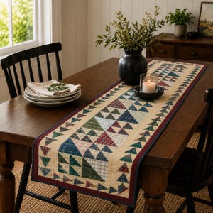 May include: A rectangular fabric table runner featuring a patchwork design of small triangles in shades of green, red, blue, and cream. The runner has a dark red border and is laid flat on a wooden table, centered with a black vase and a candle.
