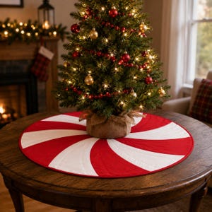 Könnte beinhalten: Ein runder Baumbehang aus Stoff mit einem rot-weißen Pfefferminz-Wirbelmuster und Steppnähten. Die Unterlage liegt flach auf einem Holztisch unter einem kleinen Weihnachtsbaum, der mit roten und goldenen Kugeln sowie Lichtern geschmückt ist.