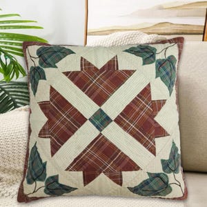 May include: Square cushion with a patchwork design. The cushion features a central starburst pattern in red and green plaid, surrounded by cream-coloured fabric and green plaid leaves. The edges are trimmed with a dark red border.