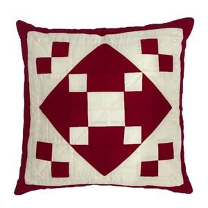May include: Square decorative pillow with a quilted design. The pillow features a central red diamond shape with white squares and a red border. The pillow is square and has a soft, inviting appearance.