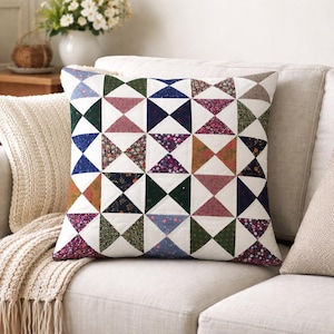 Könnte beinhalten: Quadratisches Zierkissen mit Patchwork-Design. Das Kissen zeigt ein Muster aus Dreiecken in verschiedenen Farben, darunter Blau, Grün, Rosa und Blumendrucke, vor einem weißen Hintergrund. Das Kissen liegt auf einem beigen Sofa.