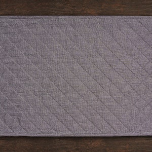 May include: A grey quilted placemat with a diamond pattern. The placemat is made of a soft, textured fabric.