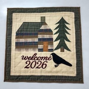 Könnte beinhalten: Eine gesteppte Wanddekoration mit einem Hütten- und Tannenbaum-Design. Die Hütte hat ein kariertes Dach und gestreifte Wände. Eine schwarze Krähe sitzt unter dem Text "welcome 2026". Der Rand ist ein Karomuster.