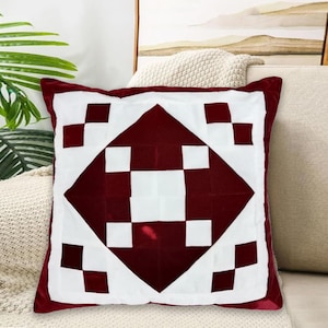 May include: Square decorative pillow with a geometric pattern in white and burgundy. The pillow has a white border and a central diamond design with squares. The pillow is on a beige sofa.