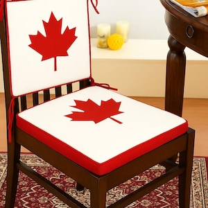 May include: A wooden chair with a white cushion featuring a red maple leaf design. The cushion has a red border and is tied to the chair with red ribbons. The chair is on a patterned rug.