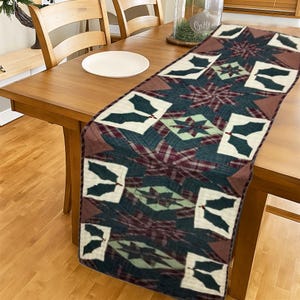 Könnte beinhalten: Ein rechteckiger Tischläufer mit Patchwork-Design. Der Läufer zeigt ein Muster aus Sternen, Stechpalmenblättern und Karos in Grün-, Burgunder- und Cremetönen. Der Läufer liegt auf einem Holztisch mit einem weißen Teller.