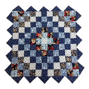 May include: Square patchwork quilt with a checkerboard pattern in shades of blue, white, and floral prints. The quilt has a dark blue zigzag border. The center features a cross design with floral fabric.