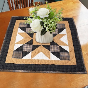 May include: A black and tan quilted table runner with a starburst design. The runner is on a wooden table with a white vase of flowers in the center.