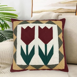 Pode incluir: Almofada decorativa quadrada com um design acolchoado com duas flores de tulipa bordô em um fundo branco. A almofada tem uma borda bordô e um padrão geométrico em tons de verde e bege.
