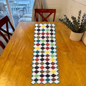 May include: A colorful diamond patterned table runner with a teal border. The runner is on a wooden table with two chairs.