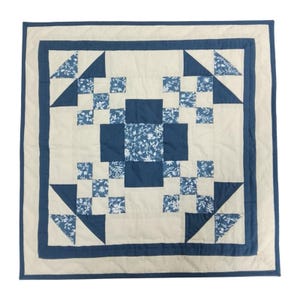 Könnte beinhalten: Ein quadratischer, handgefertigter Quilt mit einem zentralen Design aus blauen Quadraten und floralen Mustern auf cremefarbenem Hintergrund. Der Quilt hat einen dunkelblauen Rand und ist wahrscheinlich aus Baumwollstoff gefertigt. Das Gesamtdesign ist geometrisch und traditionell.